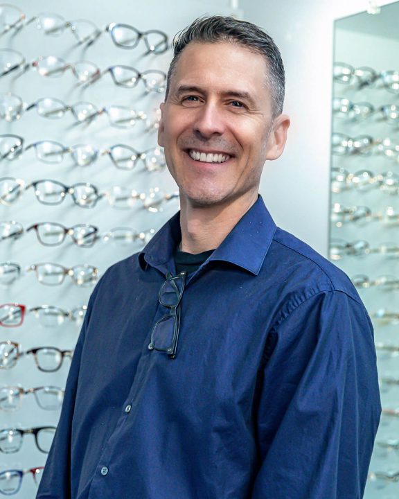 Dr Eric Lamp standing in front of a display of eyeglasses. He is wearing a blue dress shirt, standing in front of a display of glasses.