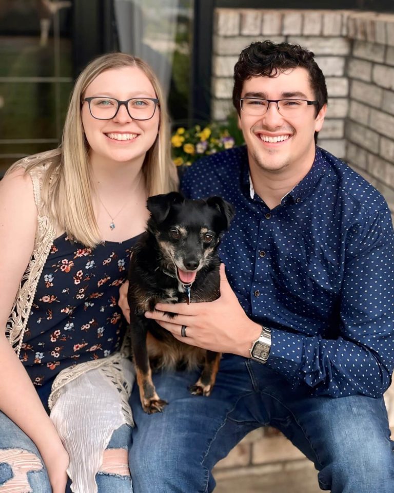 Dr. Jenna Dougherty with her husband sit smiling on a porch. They are holding a small black terrier mix.