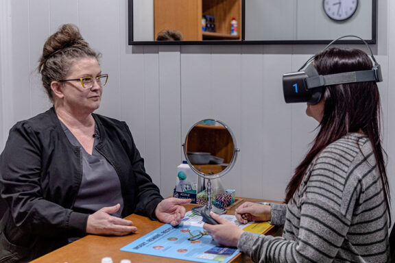 Two woman seated, one is wearing a Virtual Field headset.