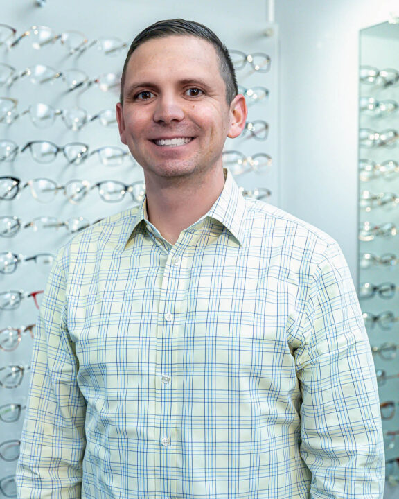 Dr. Nick Richmond standing in front of a display of eyeglasses. He is wearing a green plaid shirt and blue slacks.